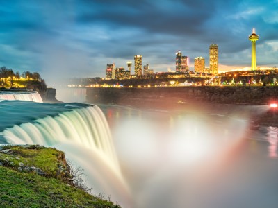 Long exposure photo of the American Falls waterfall in New York State, USA and the skyline of the city of Niagara Falls, Ontario, Canada, in the background, illuminated at twilight blue hour.